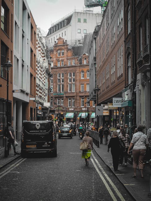 A busy urban street scene in Soho with pedestrians walking along the sidewalk and cars parked and driving on the road. The street is lined with multi-storey buildings featuring a mix of modern and historic architecture, including red brick facades and large windows. People are dressed in casual clothing, some carrying bags, and several storefronts with signage are visible on the right side. The environment appears overcast, with diffuse natural light illuminating the scene. In the background, an ornate, older building with detailed brickwork and a peaked roof stands prominently, contrasting with the surrounding contemporary structures. The image depicts a typical city street setting that could accommodate private rubbish collection services, such as Waste Disposal Soho, which specializes in alternative waste handling solutions outside conventional local authority refuse collection.