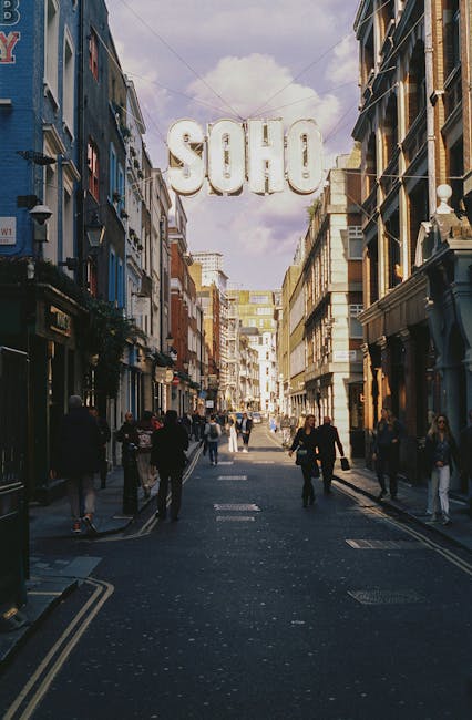 A busy urban street scene in Soho with pedestrians walking along the sidewalk and cars parked and driving on the road. The street is lined with multi-storey buildings featuring a mix of modern and historic architecture, including red brick facades and large windows. People are dressed in casual clothing, some carrying bags, and several storefronts with signage are visible on the right side. The environment appears overcast, with diffuse natural light illuminating the scene. In the background, an ornate, older building with detailed brickwork and a peaked roof stands prominently, contrasting with the surrounding contemporary structures. The image depicts a typical city street setting that could accommodate private rubbish collection services, such as Waste Disposal Soho, which specializes in alternative waste handling solutions outside conventional local authority refuse collection.