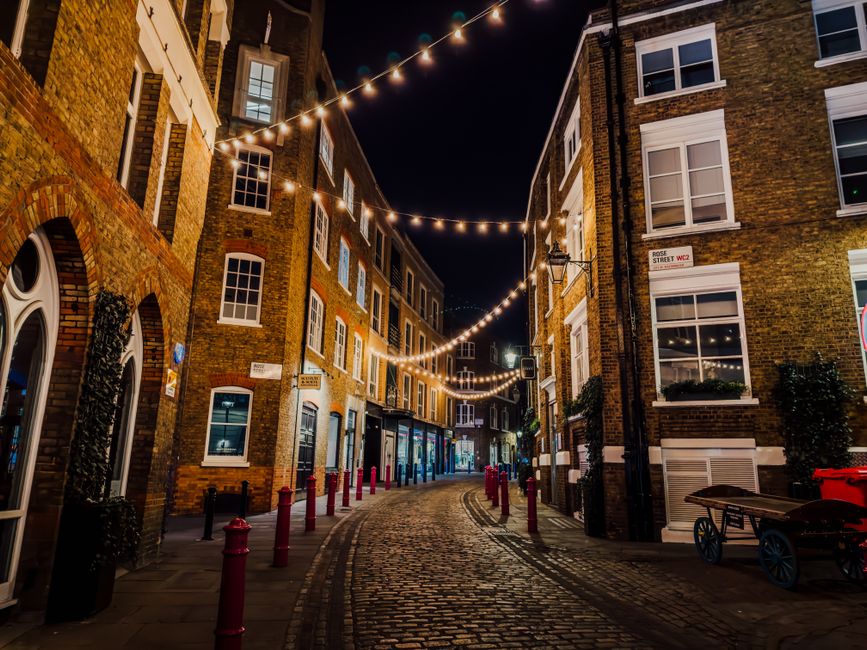 A nighttime scene of a narrow, cobblestone street in an urban area, illuminated by string lights hanging overhead between brick buildings on either side. The buildings feature a mix of traditional brick facades, with some displaying white-framed sash windows and decorative moldings. The street is lined with small bollards painted red, and a few shops or establishments with lit windows are visible along the pavement. The environment appears clean and well-maintained, reflecting a quiet atmosphere typical of an alternative waste handling or private rubbish collection setting. The lighting casts a warm glow on the textured brick surfaces and the cobblestones, emphasizing the urban character of the scene and subtly aligning with the context of independent waste management services near Soho, where on-site clearance or non-local-authority rubbish removal may be common.