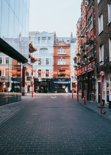 A narrow urban street scene in a city centre, lined with multi-storey buildings on both sides, featuring a mix of modern and historic architecture with brickwork and large windows. The street is paved with dark asphalt and marked with double yellow lines along the edges. Several parked vehicles, including a black van with branding for waste disposal, are positioned along the curb. Pedestrians are walking along the sidewalks, some crossing the street, and others browsing shopfronts which include cafes and retail stores with awnings and signs. Overhead, the sky appears overcast, casting diffuse natural light over the scene. The environment suggests a busy shopping district where private waste collection services, like Waste Disposal Soho, often facilitate rubbish removal for commercial premises, enabling alternative waste handling outside of local authority services.