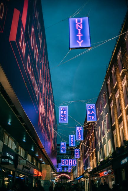 A street scene at night featuring illuminated vertical signs suspended above the pavement, with the largest sign reading 'UNITY' in bright blue letters. Other signs display words like 'STRENGTH,' 'CREATE,' 'PLANET,' 'DANCE,' 'FAMILY,' and 'BE KIND,' all in similar neon blue and pink lighting. The scene shows a lively urban environment with multi-storey buildings on either side, some with illuminated windows and brick facades. In the background, a large arched entrance is visible, contributing to the vibrant atmosphere. The street appears to have pedestrian activity, and the overall scene suggests a busy, modern area, possibly in Soho, with a focus on community and creativity. The lighting and vertical signs give a contemporary, energetic feel, which contrasts with the quieter, more utilitarian tone typical of rubbish removal services, but the detailed description of urban signage occasionally supports the visual context of on-site clearance or alternative waste handling methods at such locations.